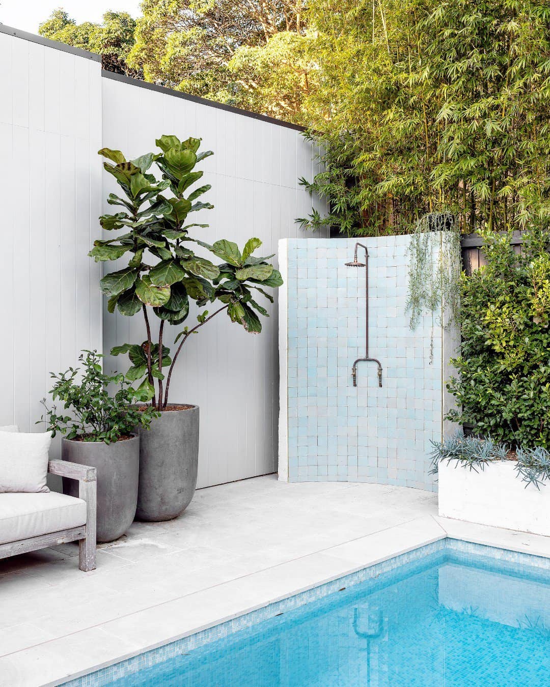 A serene outdoor shower area with white mosaic tiles next to a swimming pool, surrounded by lush greenery and two large planters with a tropical plant and a small tree, perfect for a cool-off.