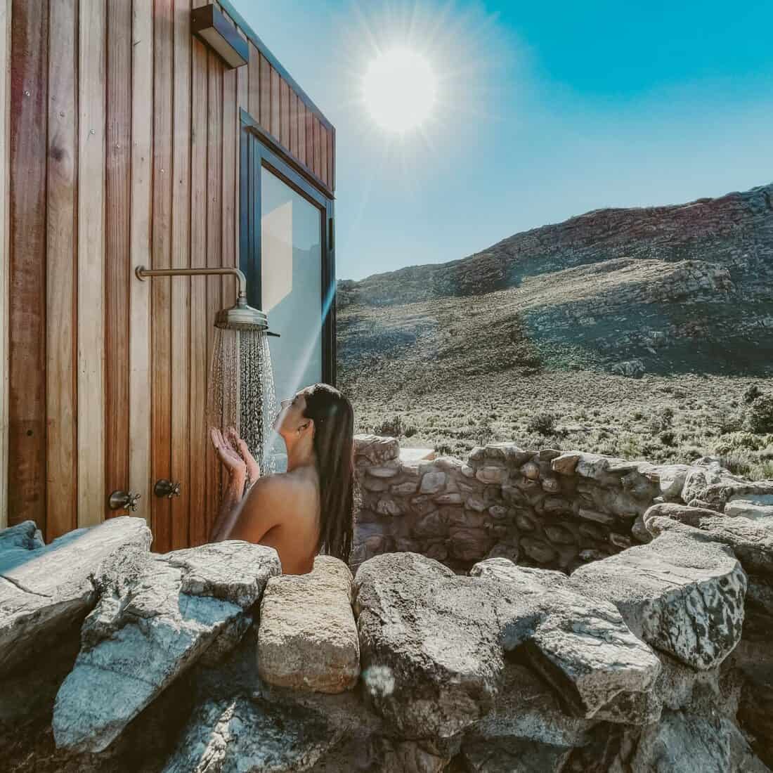 A woman enjoying a cool-off in an outdoor shower at a wooden cabin with a scenic mountain background under a clear blue sky.