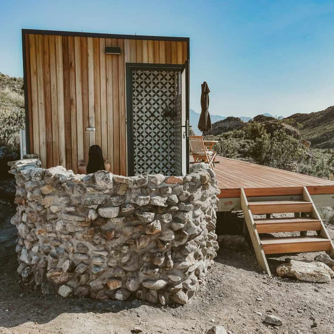 A rustic stone and wood cabin with a dark metal door, surrounded by rocky terrain under a clear blue sky, featuring outdoor shower ideas with a wooden deck and stairs on the right.