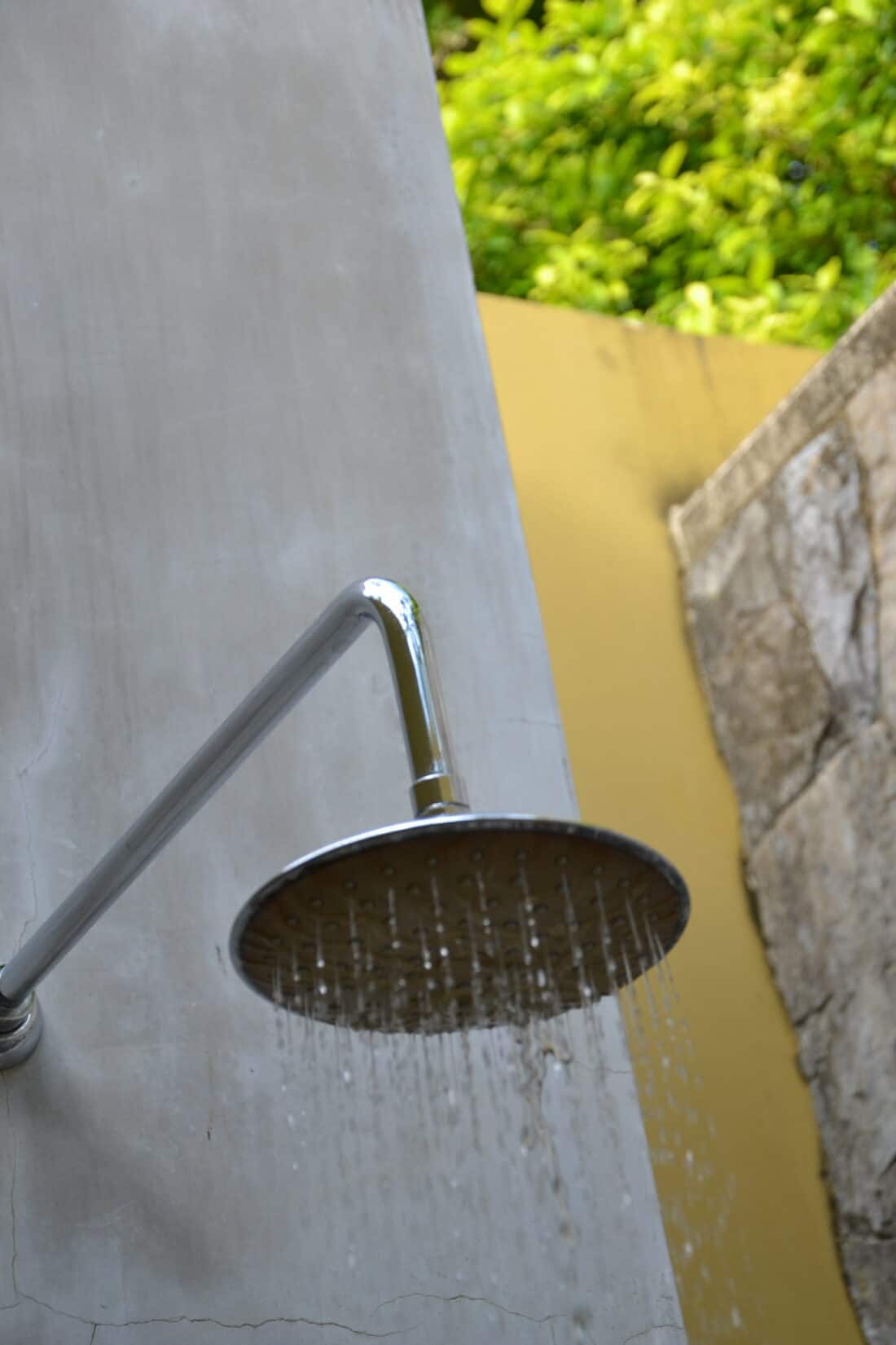 An outdoor shower head with water droplets falling, set against a blurred green and beige background, perfect for a cool-off.