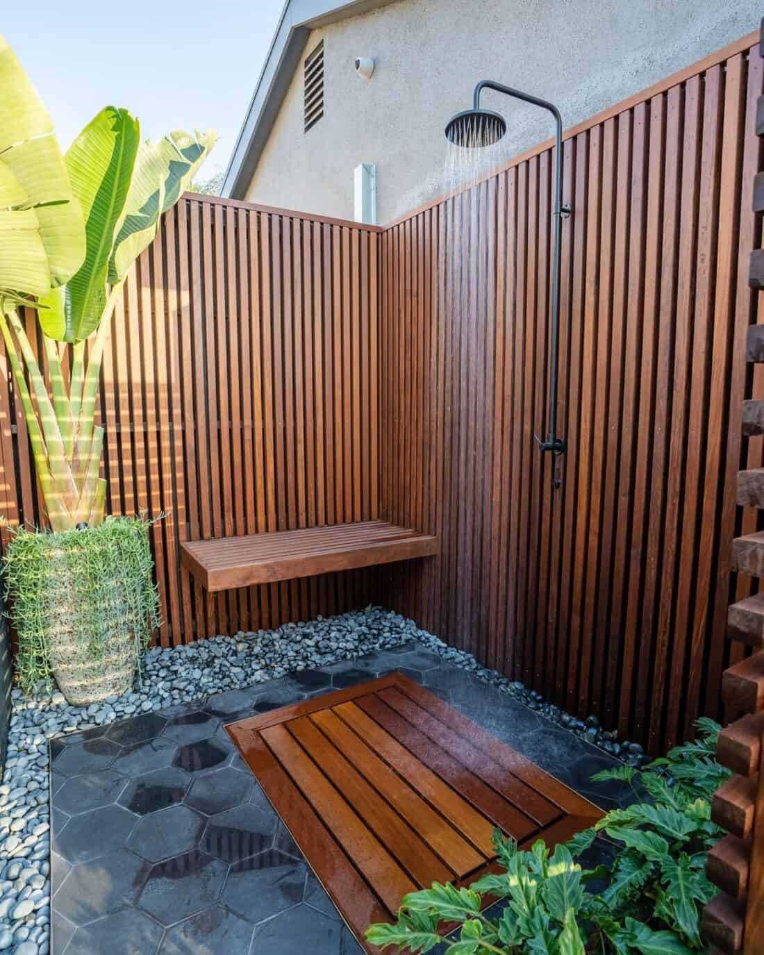 A modern outdoor shower with wooden slatted walls and flooring, featuring a black overhead shower fixture, surrounded by lush green plants and pebbles. Explore our exhaustive roundup of more outdoor shower ideas.