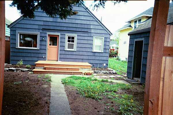 A small blue-gray house with a reddish door has a set of wooden steps leading up to the entrance. The Seattle front garden features a patchy lawn in the foreground, and a walkway leads up to the steps. A smaller matching shed is to the right before & after another house visible in the background.