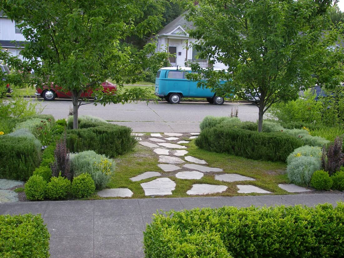 A lush Seattle Front Garden features stone pathways bordered by trimmed greenery and bushes. Two trees frame the view, leading to a street where a red car and a teal van are parked. Residential houses are visible in the background, highlighting the garden transformation that has taken place.