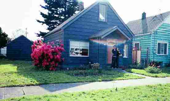 A person stands in front of a blue house with a well-kept lawn and blooming red flowers, showcasing the impressive garden transformation. Another house painted green is visible to the right. A tree stands behind the blue house, and a detached garage can be seen to the left.