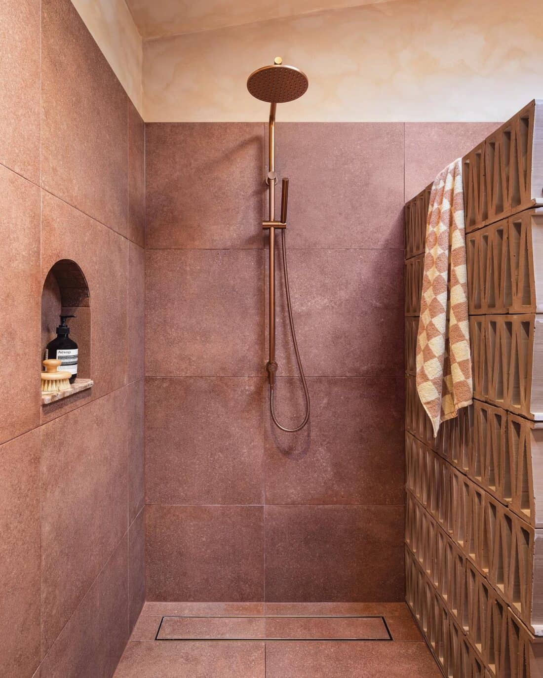 A modern outdoor shower featuring terracotta tiles with a copper rainfall shower head, a hand-held shower, and built-in shelves. A decorative towel hangs on the side.