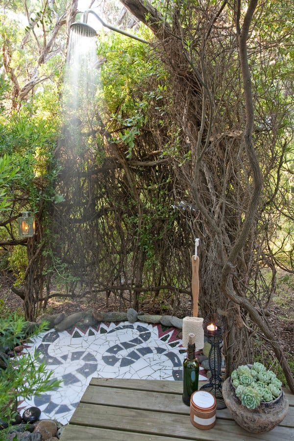 An outdoor shower surrounded by lush greenery with a geometric patterned mat on the ground and succulents decorating the wooden deck. Soft light filters through the branches, perfect for a cool-off.