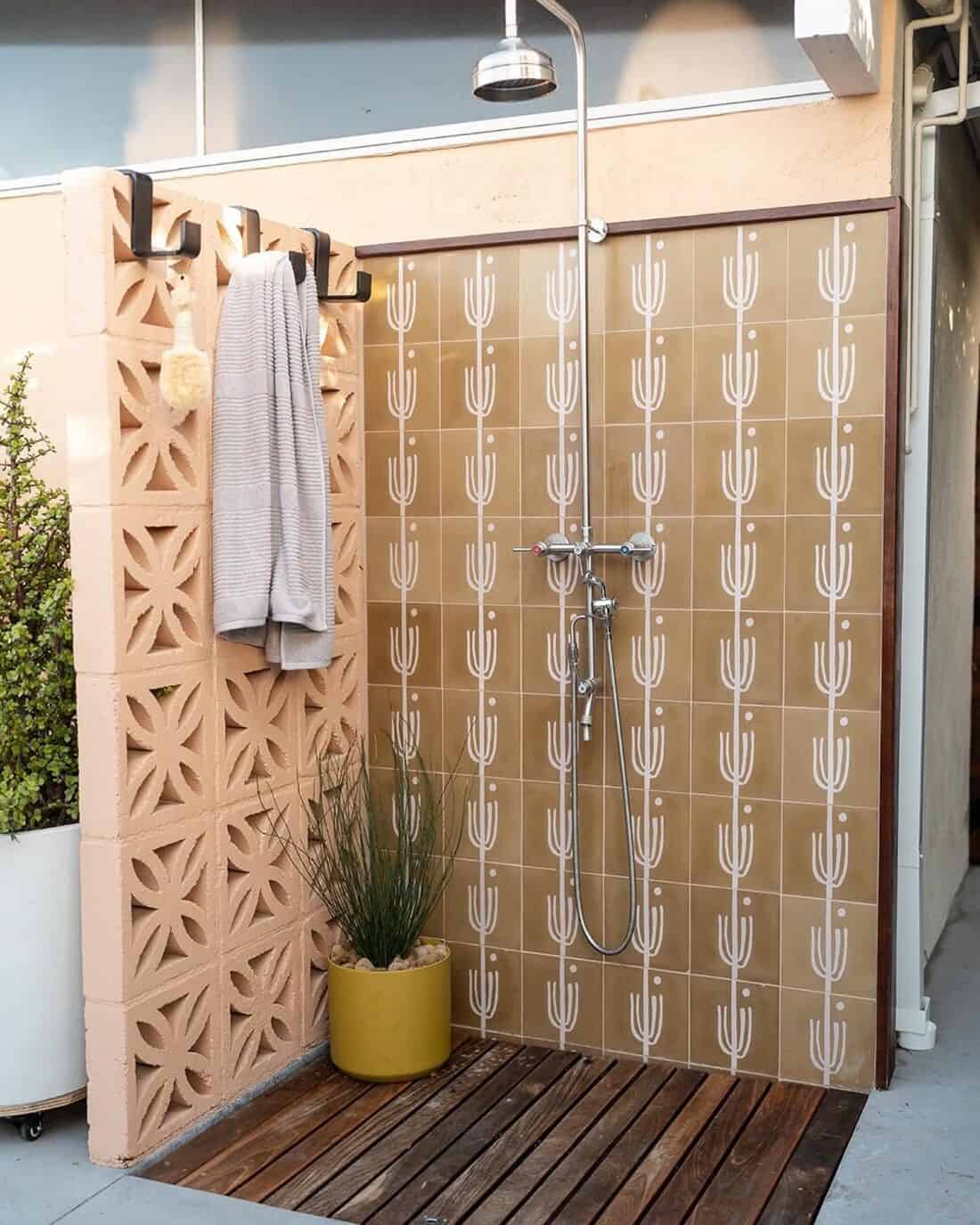 Outdoor shower setup with decorative beige tiles featuring a cactus pattern, designed for those looking to cool off, a wooden floor, a hanging towel, and a potted plant by the side.