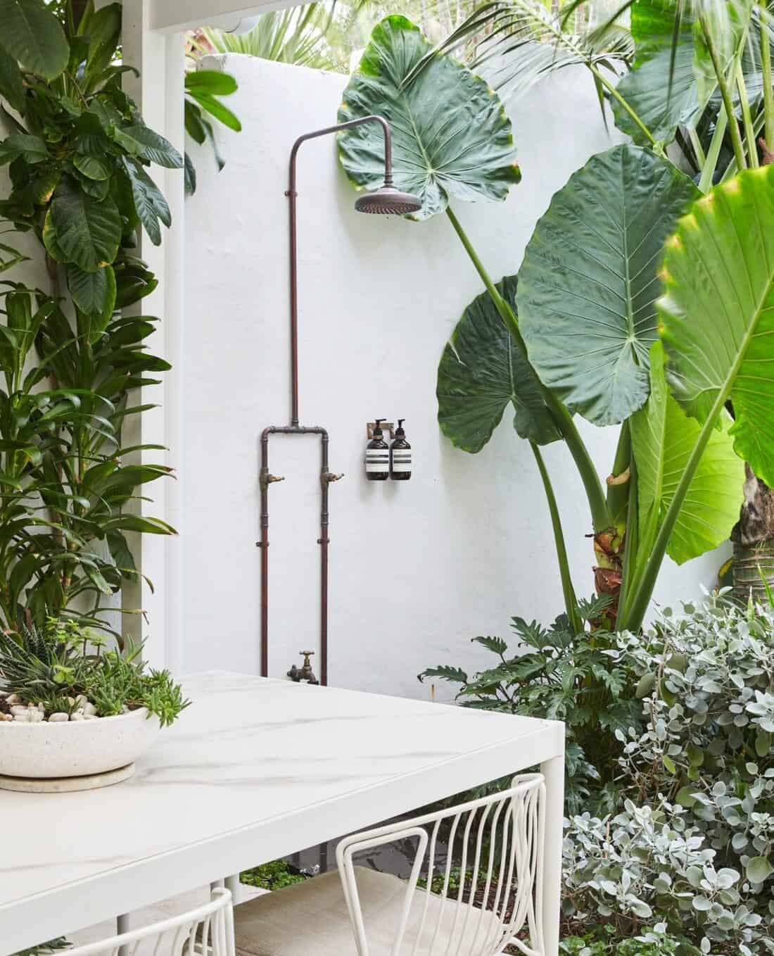 An outdoor shower surrounded by lush tropical plants, providing cool-off relief. This shower features a vintage-style overhead head and dark metal piping, alongside a white table and chair.