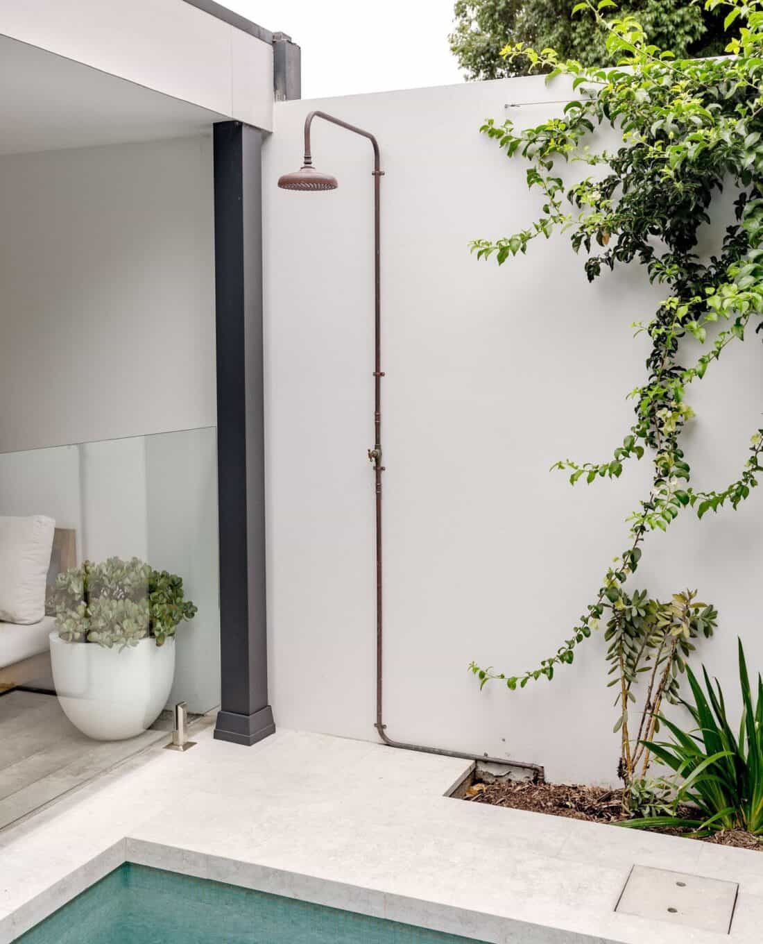 An outdoor shower area next to a small pool, featuring a wall-mounted showerhead, plants in pots, and clean, minimalist design with grey and white tones. Ideal for a cool-off during hot days
