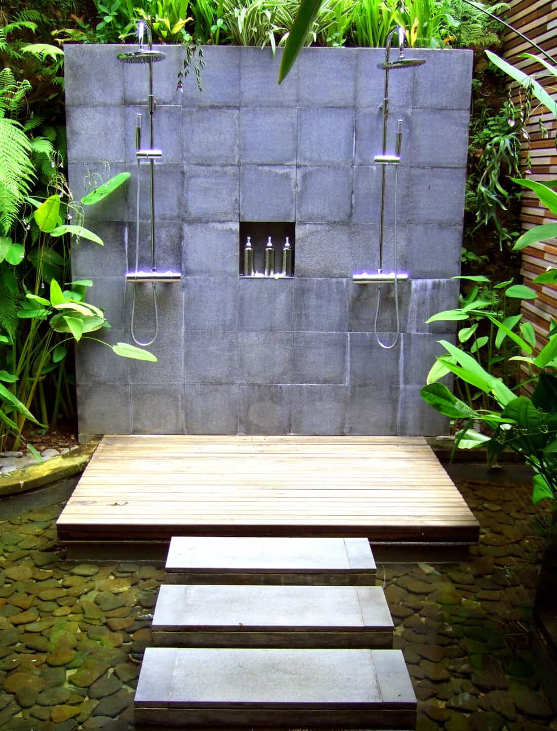 Outdoor shower with dual heads on a gray concrete wall, a wooden platform in front, and stepping stones across the water, nestled amid lush greenery.