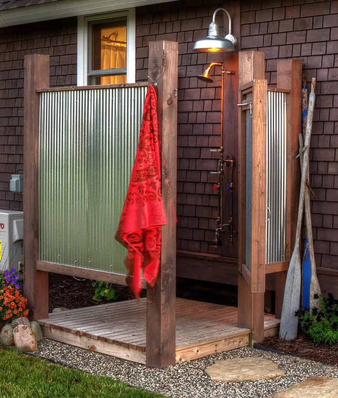 An outdoor shower with a red towel hanging on a wooden frame, a rustic door partially open, and garden flowers nearby, adjacent to a house with brown siding offers a great way to cool-off.