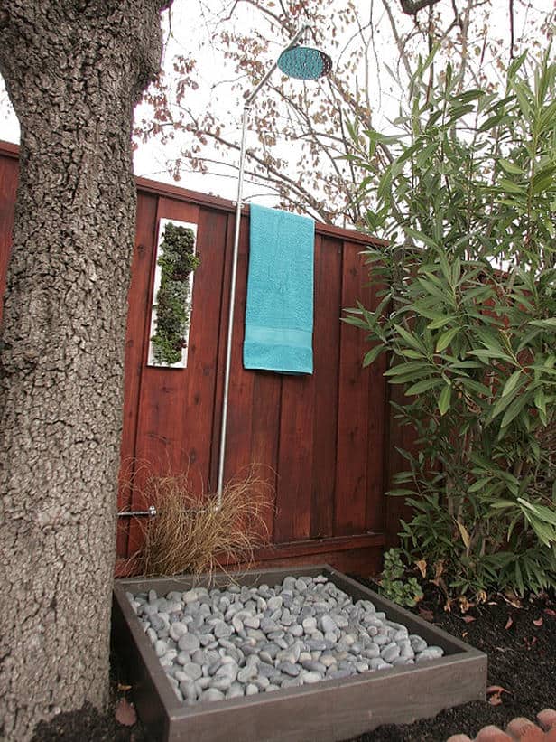 A serene backyard corner featuring a wooden fence, a textured tree trunk, an outdoor shower with a suspended blue towel, and a shallow box filled with smooth grey stones beside green foliage.