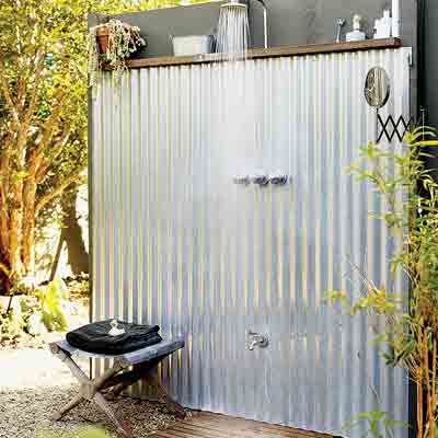An outdoor shower made of corrugated metal with a rustic wooden stool and a small black bag beside it, set in a garden setting with lush greenery, provides cool-off options in this exhaustive roundup