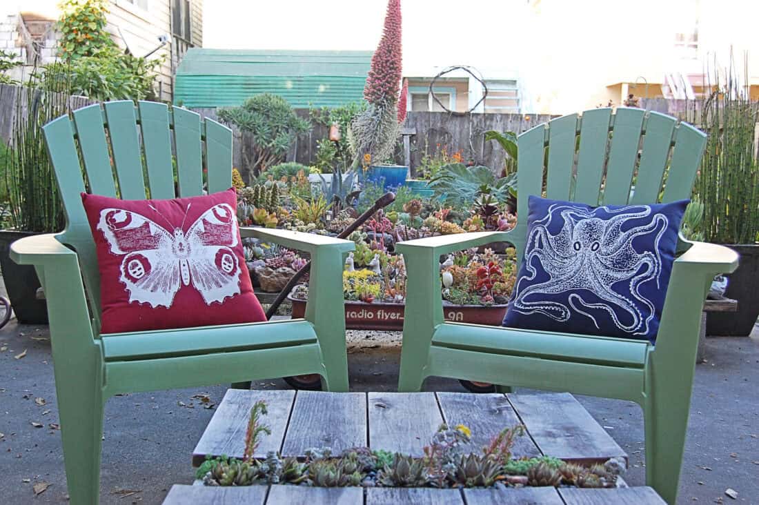 Two green Adirondack chairs with red and blue decorative pillows face each other, flanking a small wooden table with plants, in a succulent garden.