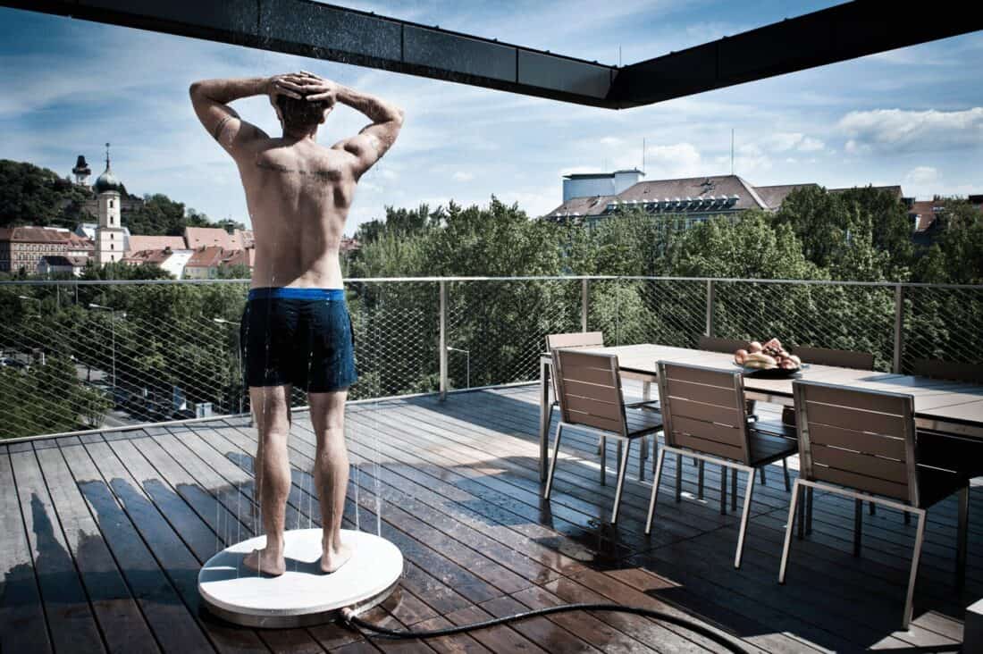 A man standing on an outdoor terrace, holding his head with both hands, facing a scenic city view. He wears blue swim shorts, indicative of preparing to cool-off, with patio furniture and greenery