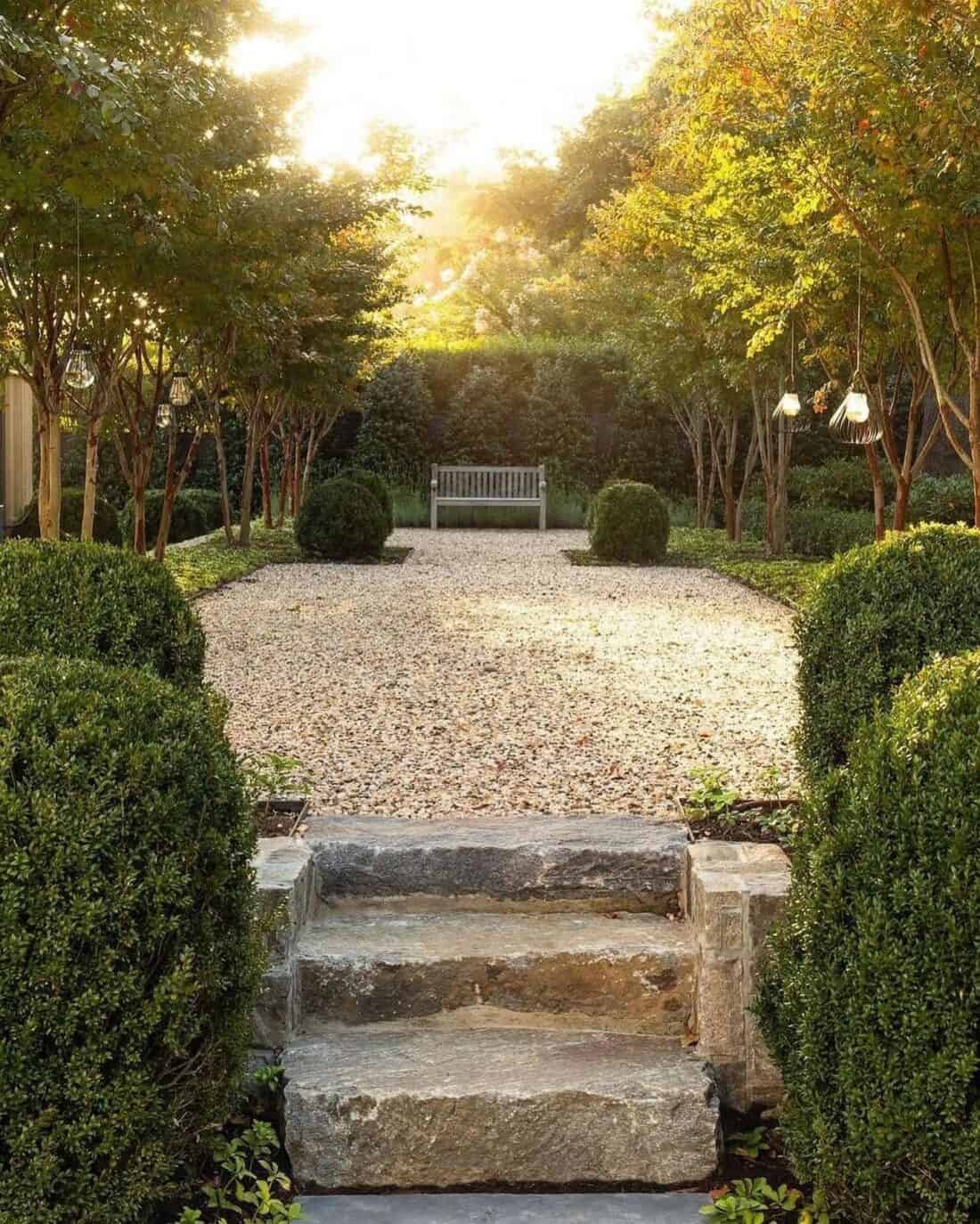 A serene garden scene with a gravel pathway leading to a wooden bench bathed in sunlight. Stone steps in the foreground are bordered by neatly trimmed hedges and trees adorned with hanging lanterns, perfect for transforming into a backyard yoga space. Lush greenery surrounds the area, creating a peaceful ambiance.