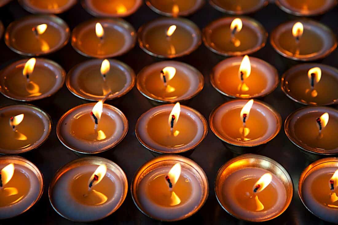 A collection of lit candles in small, round metal holders are arranged in neat rows, illuminating a backyard yoga space. The candles emit a warm, glowing light, creating an atmosphere of peace and tranquility. The background is dark, emphasizing the brightness of the flames.