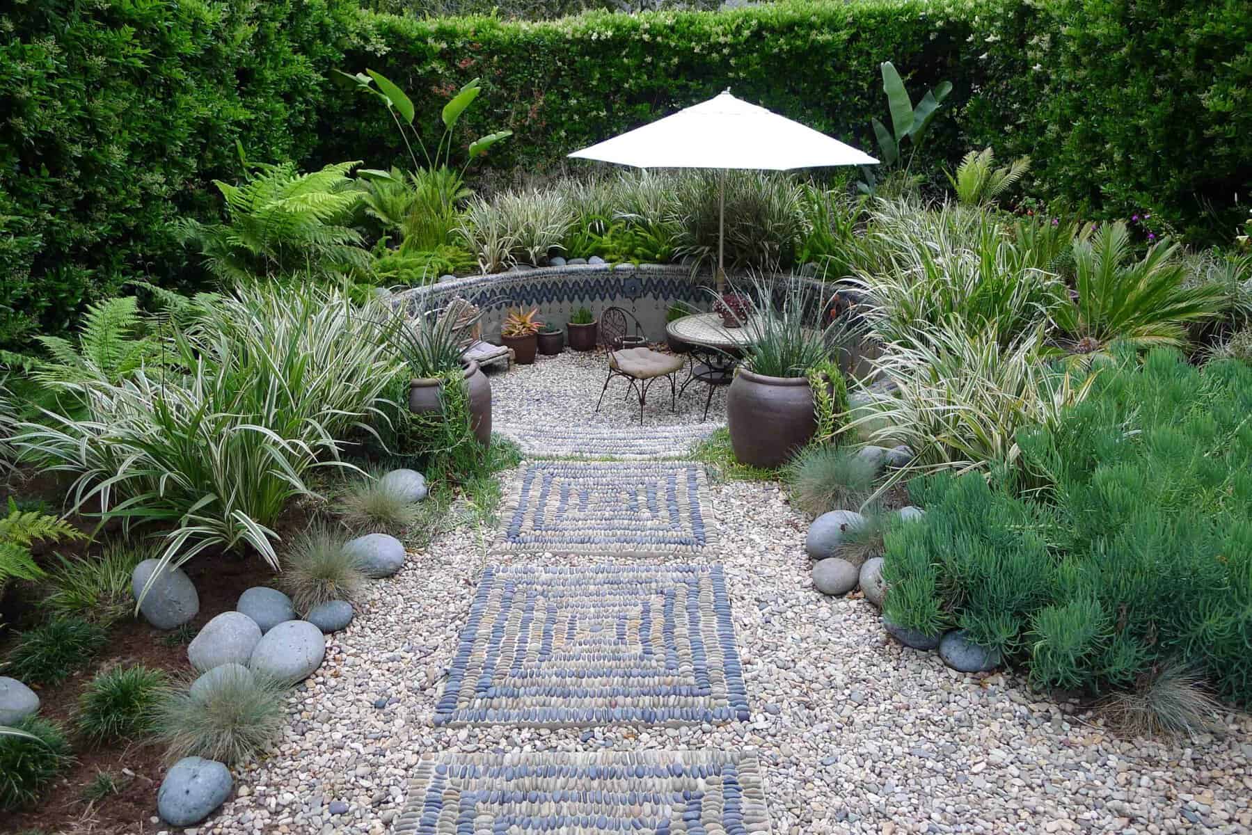 A lush garden features a stone pathway leading to a cozy seating area with a round table, two chairs, and a white umbrella. Greenery, including various shrubs and tall plants, surrounds the space. Several large potted plants and decorative stones complete this ideal backyard yoga studio.