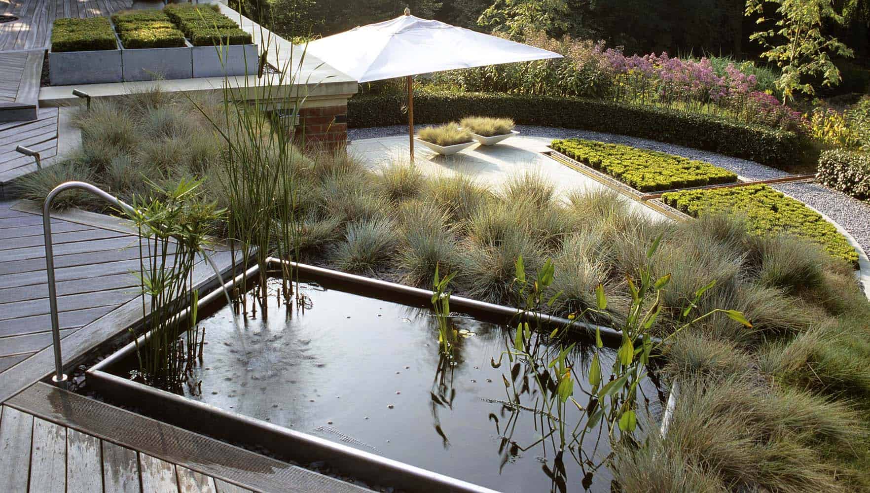 A serene rooftop garden, showcasing garden design basics, features a rectangular reflective pond with aquatic plants, surrounded by tall grasses. Adjacent is a wooden deck leading to a sleek staircase. A white umbrella shades a small seating area next to another landscaped section with lush greenery.