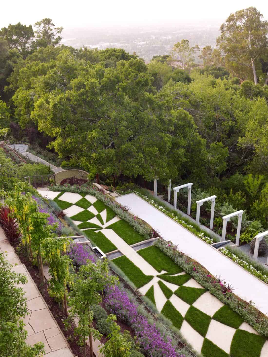 A beautifully landscaped garden with patterned lawn areas and purple flowers. A pathway with white archways leads through the Garden Gallery. Dense green trees and a distant Los Gatos cityscape are visible in the background, while greenery lines the path and garden edges.