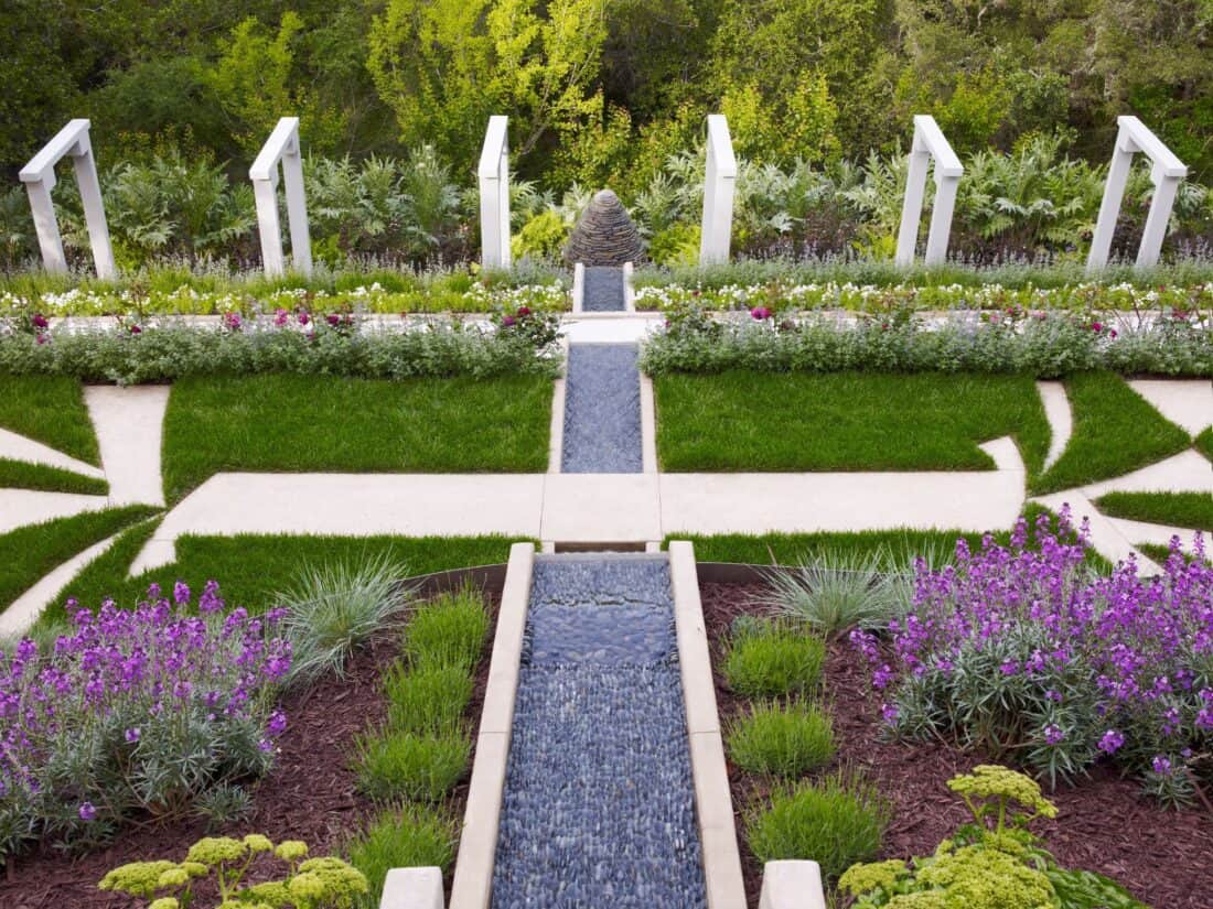 A well-manicured garden in Los Gatos featuring geometric pathways, lush green grass, and vibrant flower beds. A blue stone water feature runs through the center, flanked by white columns and surrounded by various shrubs and blooming purple and yellow flowers—a true Garden Gallery.