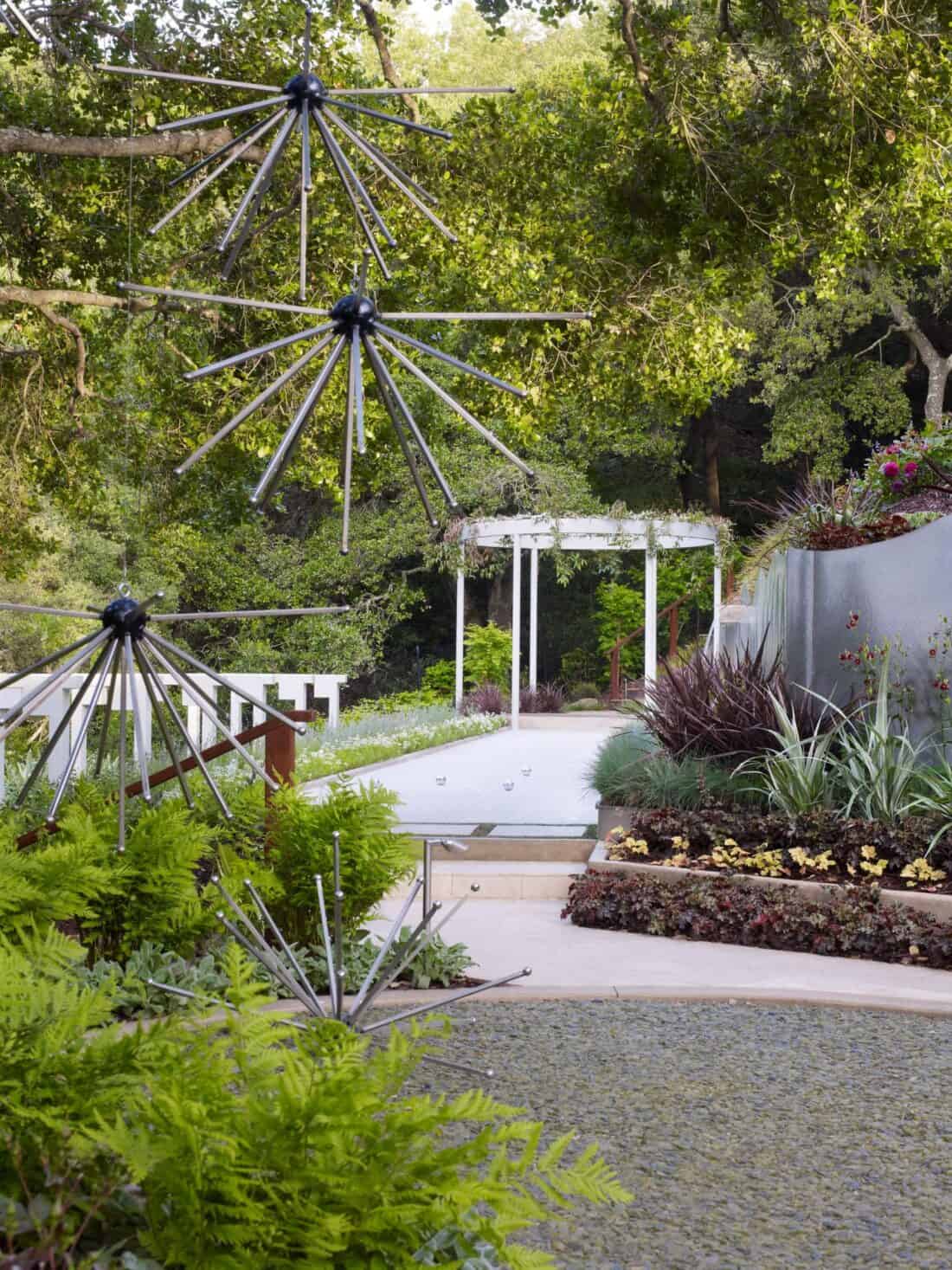         A tranquil garden pathway in the Garden Gallery of Los Gatos features modern, starburst metal sculptures hanging and placed on the ground. The path leads to a white pergola surrounded by lush greenery and a variety of plants, including ferns and succulents. Trees and bushes create a serene backdrop.