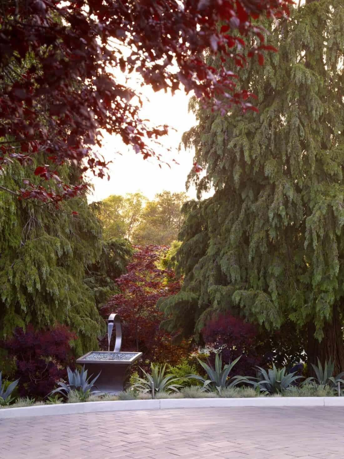 A tranquil garden scene at the Garden Gallery in Los Gatos features a sundial sculpture surrounded by lush greenery, tall trees, and vibrant plants. The sun's warm light filters through the canopy, creating a serene and inviting atmosphere.