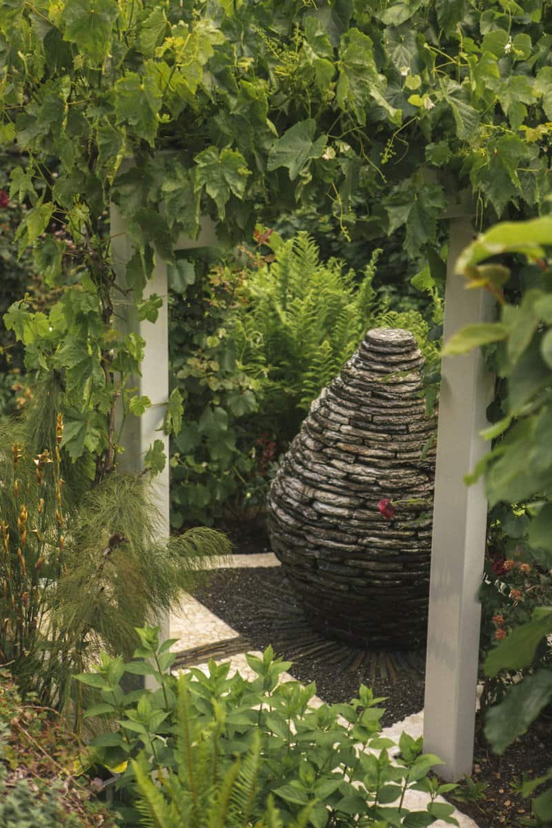 A stone sculpture resembling a beehive is surrounded by lush greenery in Los Gatos Garden Gallery. The sculpture is sheltered by a white wooden pergola, with various plants and vines growing around it.