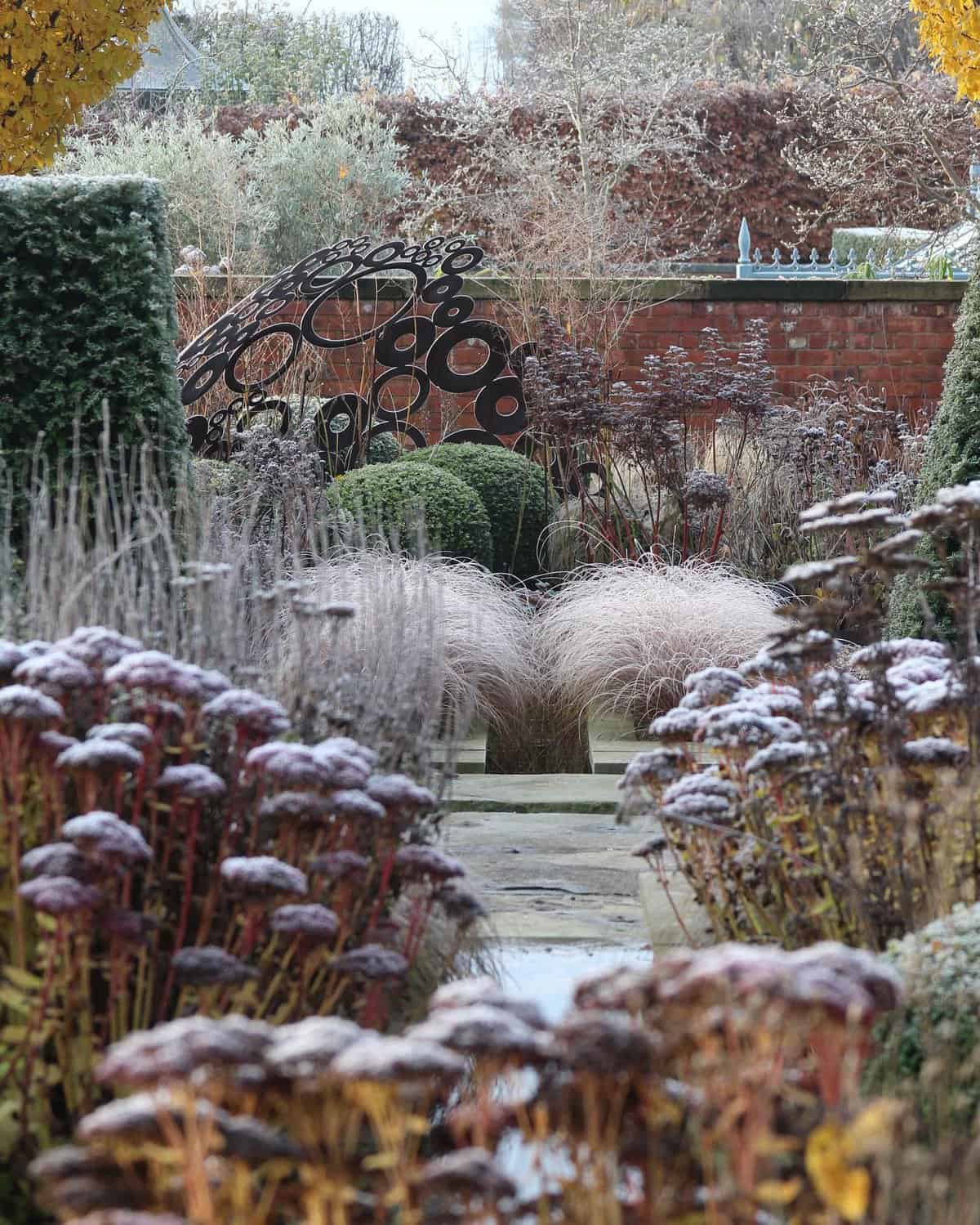 A winter garden scene featuring frost-covered plants and shrubs evokes a serene transition from an autumn garden. Ornamental grasses and sedum line a stone pathway leading to a metal sculpture with circular designs, while a red brick wall and leafless trees form the backdrop.