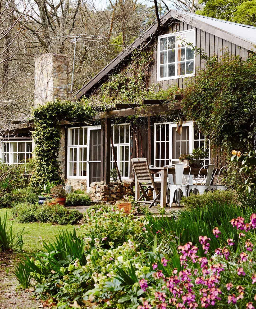 A quaint, charming cottage with ivy-covered walls and a stone chimney. The front yard, inspired by Edna Walling's garden design, is lush with greenery, pink flowers, and shrubs. There's an outdoor seating area with a table and chairs on a stone-paved patio. Trees surround the serene setting.