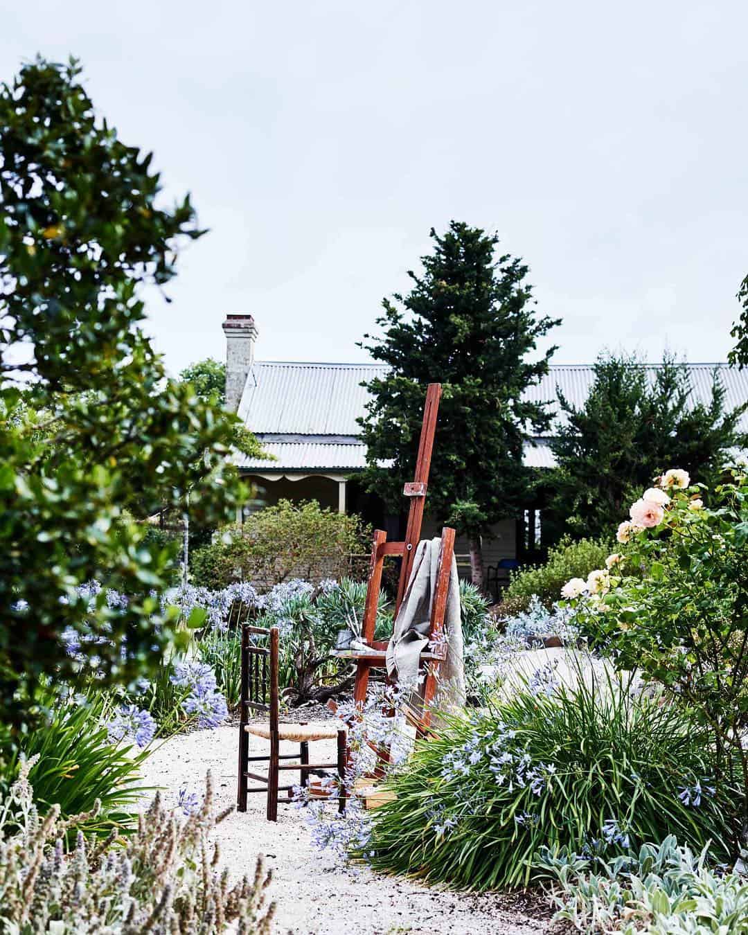 A tranquil garden scene with a wooden easel and chair on a gravel path reflects Edna Walling's garden design principles. The easel holds a canvas covered with a cloth, surrounded by various plants and flowers. A house with a corrugated metal roof is partially visible in the background.