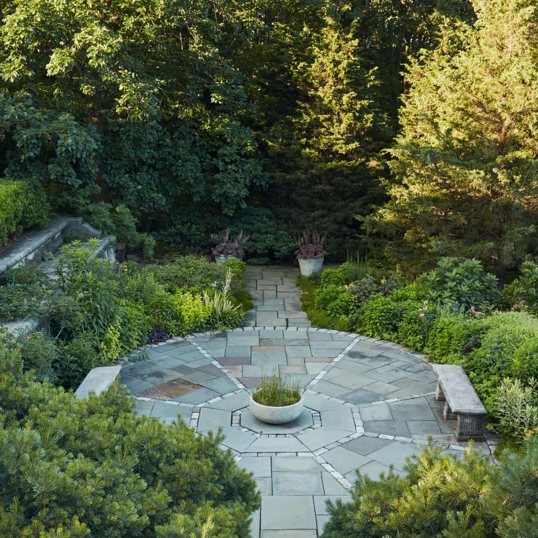 A peaceful garden features a circular stone patio with a central planter. Two stone benches are situated on either side, surrounded by lush greenery, trees, and various plants, including ilex verticillata. Steps bordered by hedges lead to the patio area from the left side.