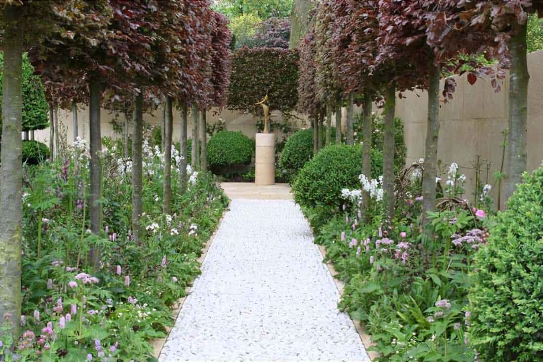 A white pebble pathway flanked by lush, green aerial hedges and flowering plants leads to a focal point - a statue atop a stone pedestal. Tall trees with sculpted branches create a canopy over the garden path.