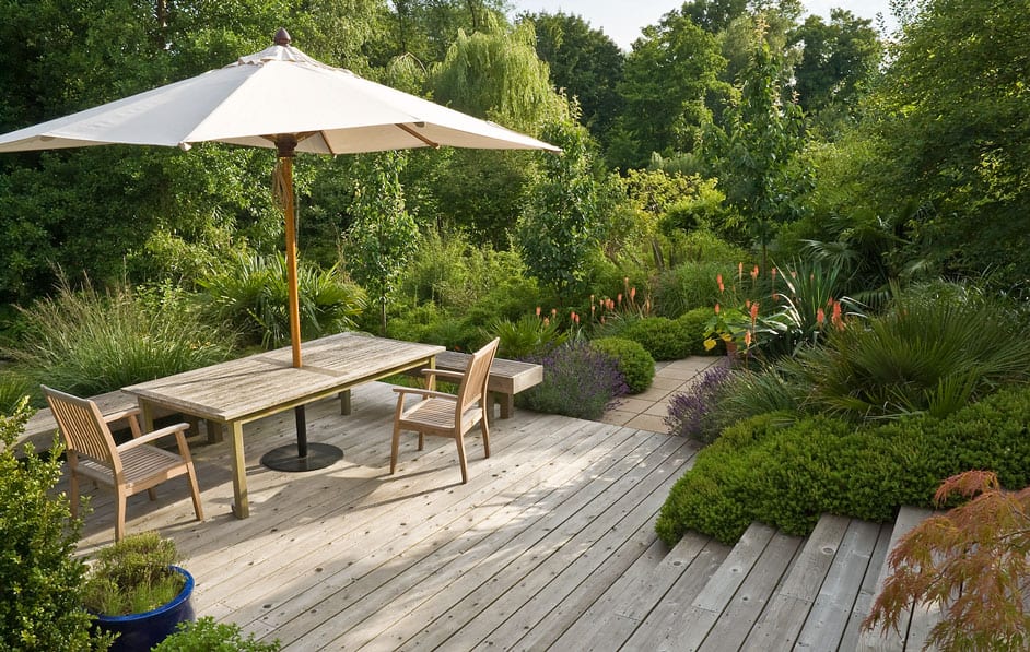 A wooden deck with a large umbrella shading a table and four chairs, surrounded by lush green foliage and various garden plants, including bushes and flowers. Steps lead down from the deck to a garden path in this serene and inviting scene at Acres Wild.