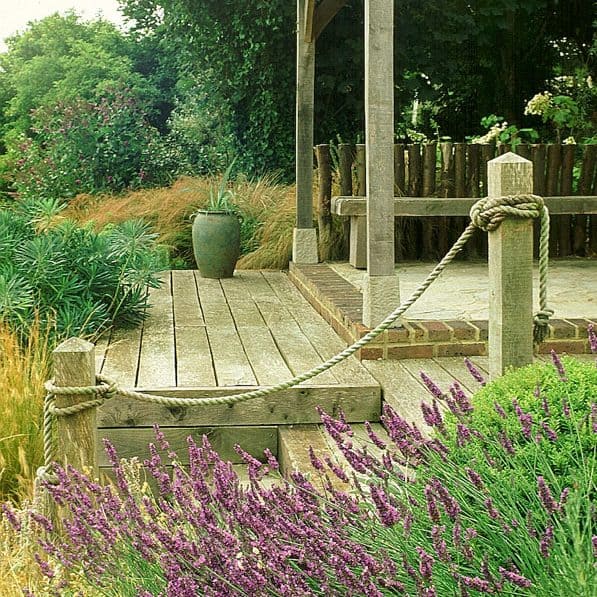 A serene garden scene with a wooden patio and rope railing, surrounded by lush greenery and purple lavender flowers in the foreground. A large green planter sits on the patio, and an overhanging pergola provides shade. Trees and shrubs fill the background, creating a Daily Garden haven.