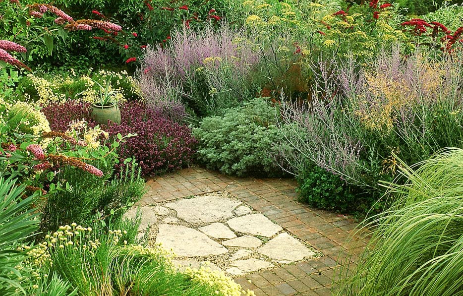 A beautifully landscaped Wild & Windswept Garden featuring a brick and stone patio surrounded by lush greenery and blooming flowers in various colors, including purple, yellow, and red. The area exudes serenity and vibrant natural beauty.