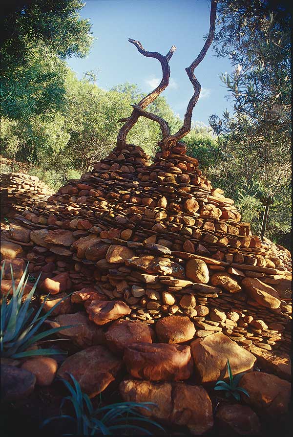 Cairn made of flat stones piled high with two twisting tree branches on top, nestled in a verdant South African forested area. The clear and sunny sky casts warm light on the rockery, illuminating the surrounding greenery and trees.