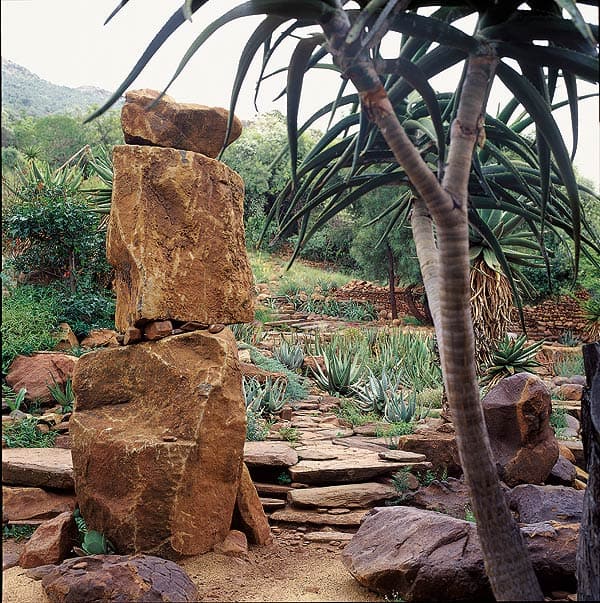 A natural garden scene features a large rockery with stacked rock formations in the foreground and lush greenery throughout. Various succulents and cacti are scattered around, complemented by tall South African agave plants. A stone pathway meanders through the garden.