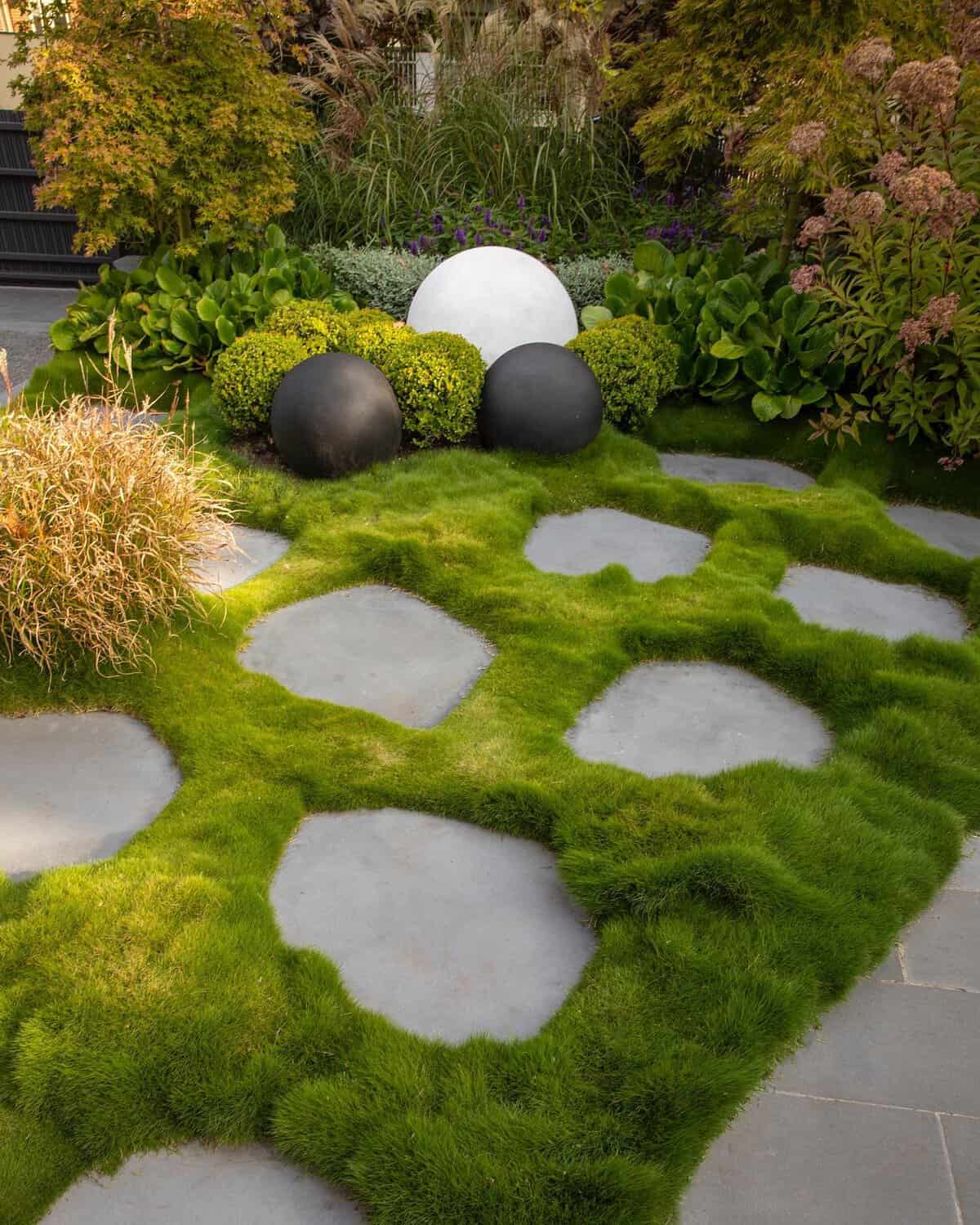 A modern garden features irregularly shaped stepping stones embedded in a lush Korean No-Mow Grass. Decorative black and white spheres adorn a neatly trimmed bush area in the background, surrounded by various plants and greenery.