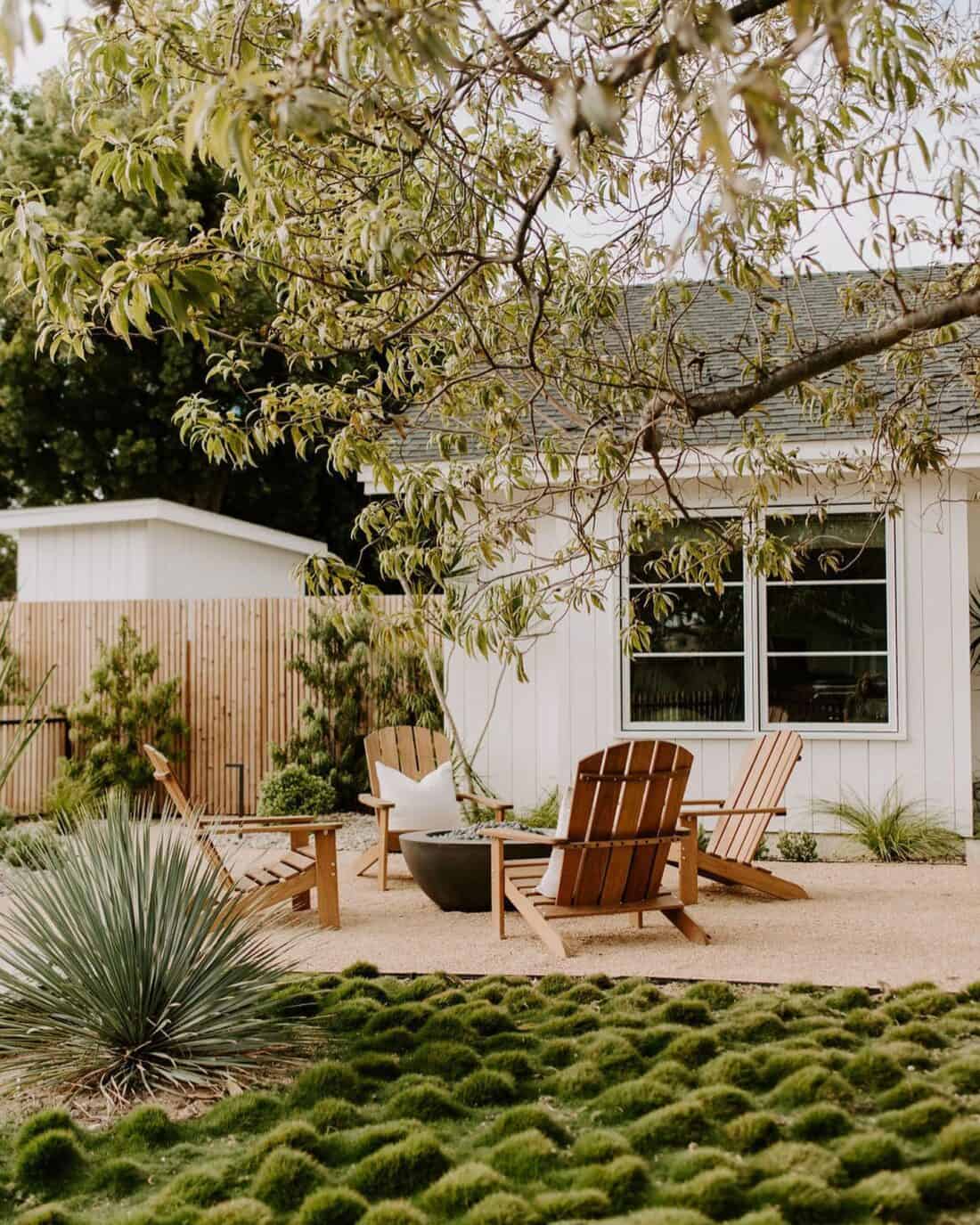 A tranquil backyard featuring wooden Adirondack chairs around a modern fire pit on a gravel surface. Lush greenery, including Korean No-Mow Grass and a leafy tree, dot the landscape. A white house with large windows and a wooden fence provide a serene backdrop.