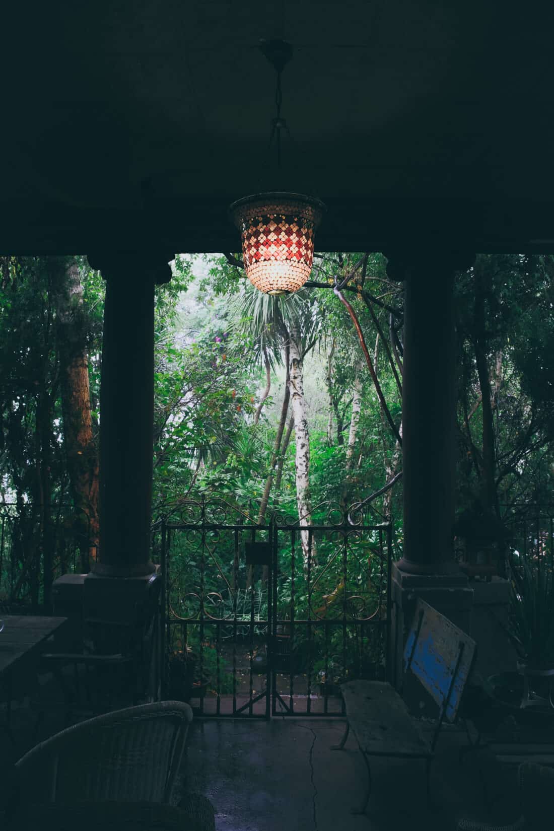 A dimly lit porch featuring a neglected hanging chandelier with red and white patterns. In the background, lush green foliage of a Victorian garden is visible through an ornate metal gate flanked by large pillars. A bench and wicker furniture are situated on the porch area.