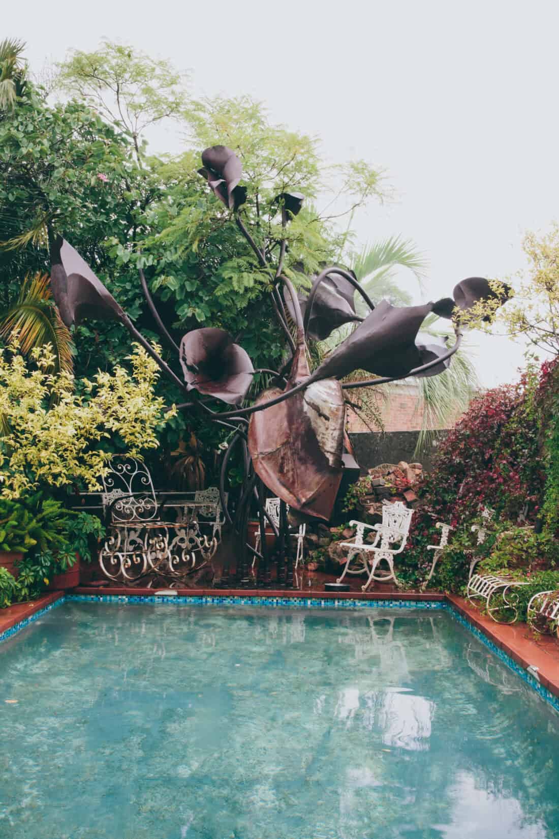 A serene poolside garden featuring a clear swimming pool, surrounded by vibrant greenery and lush plants. A large metal flower sculpture stands prominently, and white Victorian-style garden furniture, including chairs and a table, adds a touch of elegance to the setting.