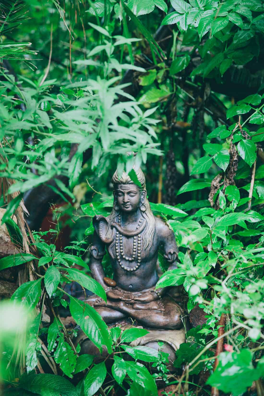 A statute of a meditative figure with long hair and intricate adornments sits surrounded by lush green foliage in a neglected Victorian garden. The serene statue appears to be made of bronze and is partially covered by natural growth, creating a peaceful, nature-integrated scene.