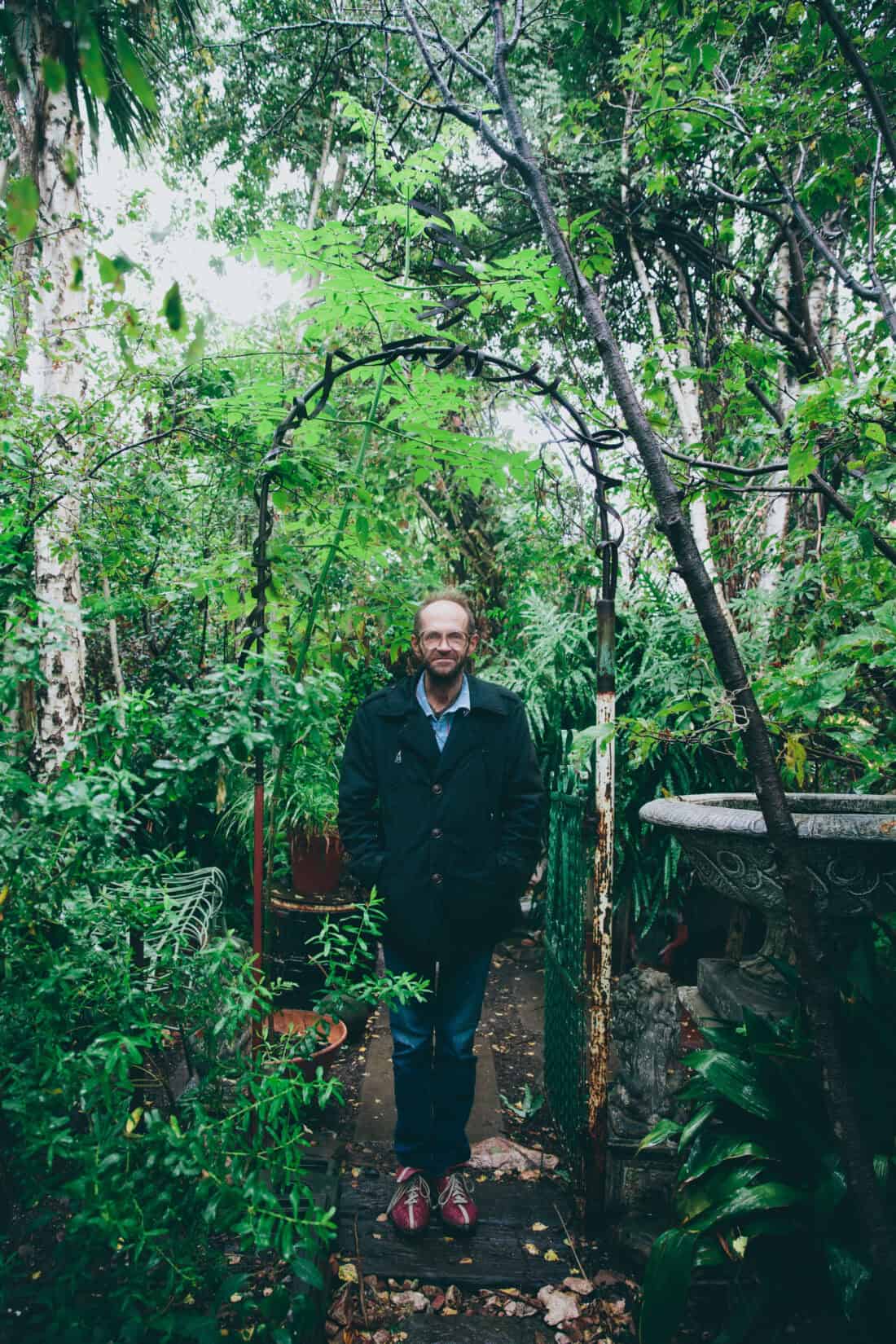 Clive Rundels, a person with glasses and a beard, stands in the middle of a lush, green garden filled with various trees and plants. They are wearing a dark jacket, jeans, and red shoes. An archway made of branches and a birdbath grace this seemingly neglected Victorian garden in the background.