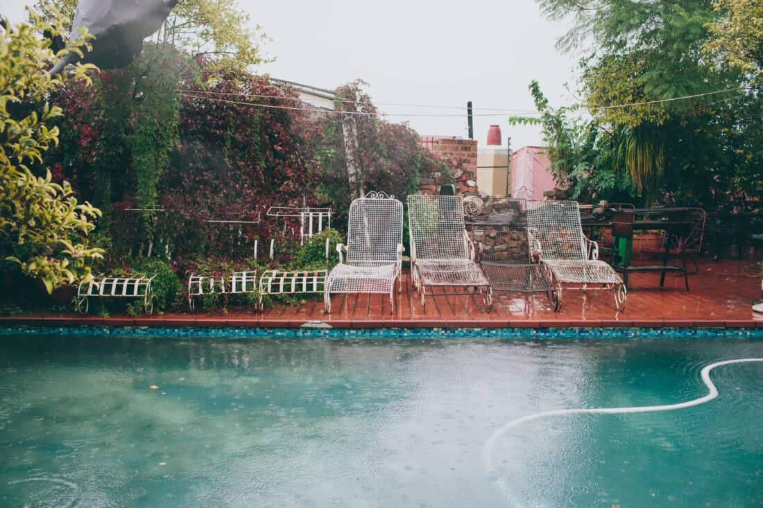 A swimming pool with raindrops on the water surface, bordered by a brick patio area. Several metal lounge chairs and a table set are arranged on the patio. Green trees and climbing plants surround the area, reminiscent of a neglected Victorian garden, creating a lush atmosphere.