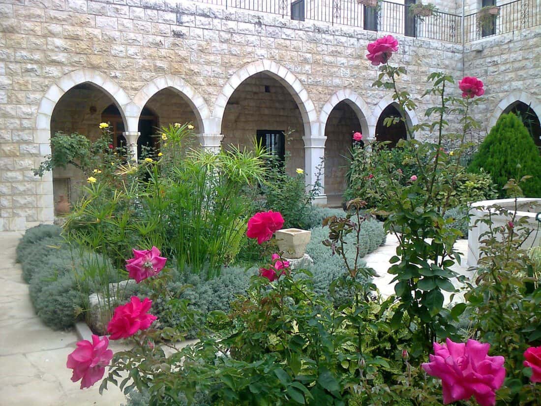 A serene courtyard garden with stone arches in the background captures the Romance of the Rose. The garden features lush green foliage, bright pink roses in the foreground, and various plants surrounding a small grassy area. The stone building, reminiscent of Pat Leuchtman's designs, boasts multiple arches and balconies.