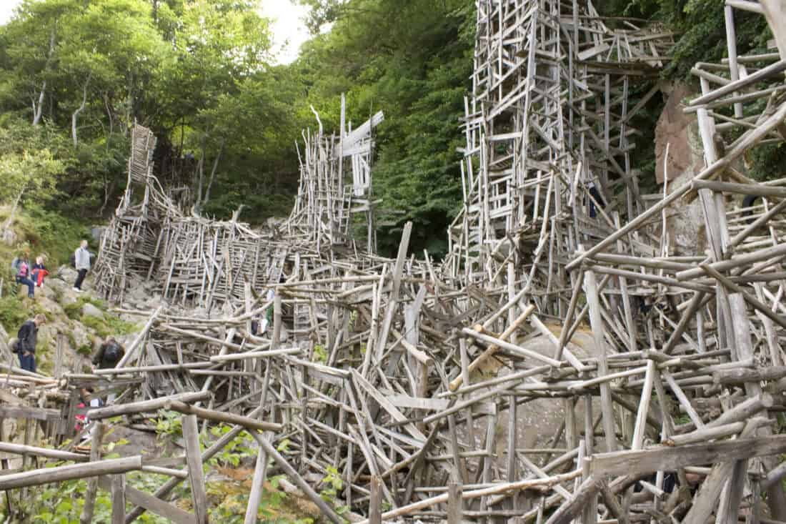 An intricate, large-scale wooden structure composed of interwoven driftwood and planks stands in a forested area. Tourists are navigating the pathways within the material-rich structure, and lush green trees are visible in the background.