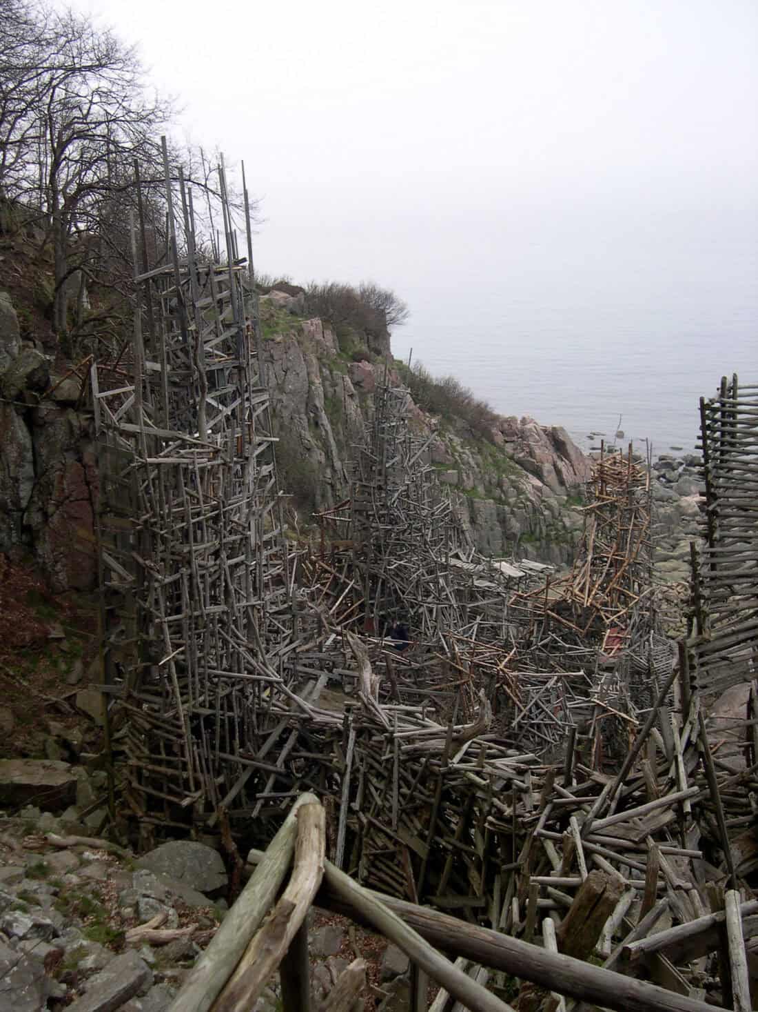 An intricate wooden structure of interlaced driftwood sticks and branches stands on a rocky coastal hillside. The tall, spindly construction appears artistic, with multiple towers reaching towards the sky. Set against a foggy seascape backdrop, it could serve as an enchanting garden centerpiece.