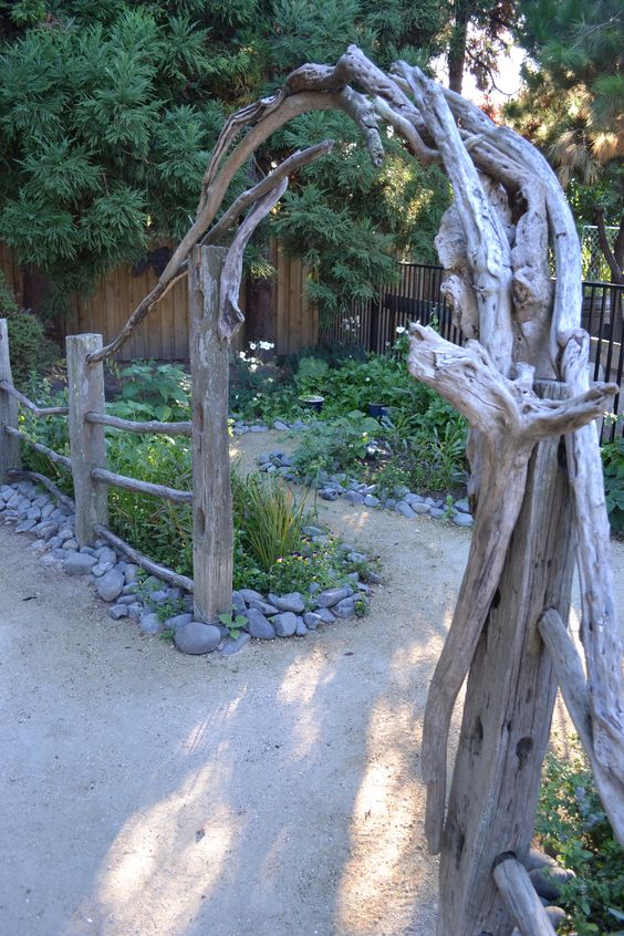 A rustic garden pathway featuring a wooden trellis made of intertwined, weathered driftwood branches forming an arch. The path is lined with stone borders and surrounded by greenery, flowers, and a wooden fence in the background. Pine trees add a lush backdrop, showcasing creative garden uses of natural materials.