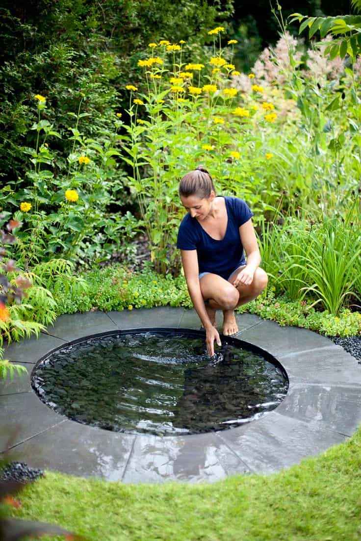 A woman in a navy blue shirt and shorts kneels beside a small round garden reflecting pool with a stone border in a lush garden. She bends over to touch the water mirror with her hand, surrounded by green plants and tall yellow flowers in full bloom.